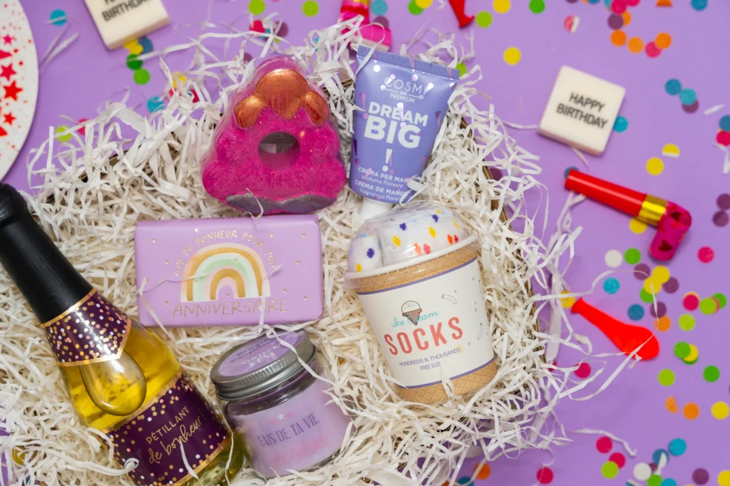 Contenu d'une box cadeau d'anniversaire surprise avec bouteille de pétillant, bougie, produits cosmétiques, chaussettes et confettis sur fond violet.