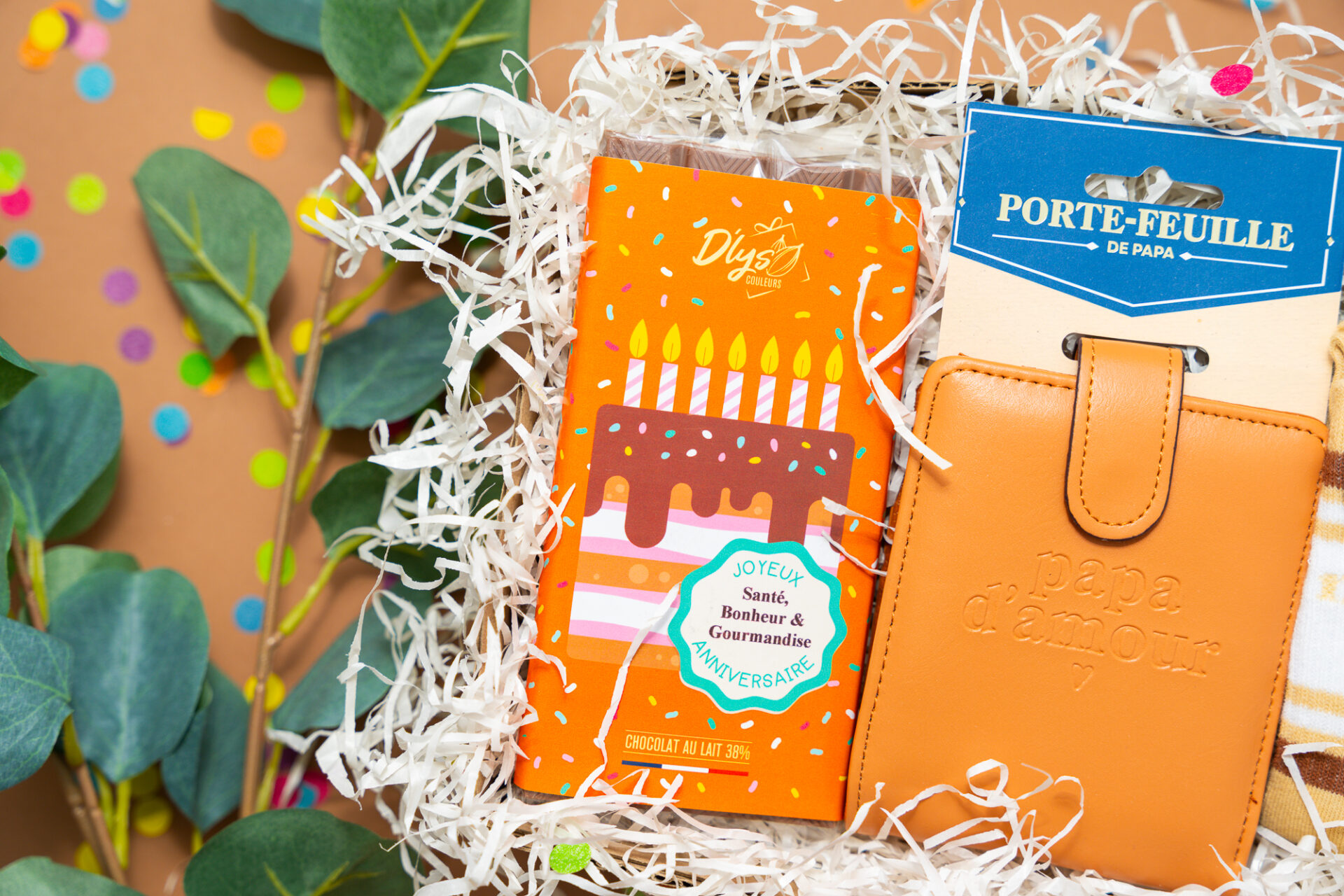 Vue aérienne d'un coffret cadeau d'anniversaire garni d'une tablette de chocolat festive, d'un portefeuille marron et de chaussettes à rayures entourés de feuillage.