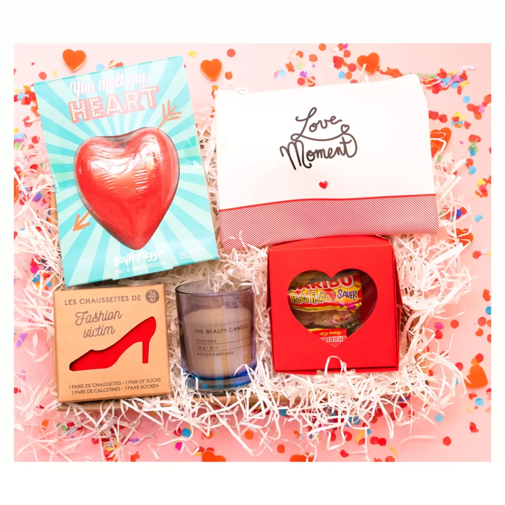 Coffret cadeau mignon pour femme contenant une trousse Love, des friandises, une bombe de bain cœur, une bougie et des chaussettes sur fond de confettis.