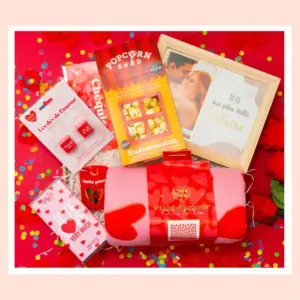 un coffret cadeau romantique “Pop The Date” posé sur un fond rose avec confettis, montrant plusieurs éléments : une boîte de popcorn gourmet, un cadre photo avec un message romantique, un plaid rose à motifs de cœur, une tablette de chocolat au design “I Love You” (ou message similaire), un petit jeu de dés “Les dés de l’amour”, et d’autres petites surprises d’ambiance Saint-Valentin.