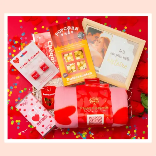 un coffret cadeau romantique “Pop The Date” posé sur un fond rose avec confettis, montrant plusieurs éléments : une boîte de popcorn gourmet, un cadre photo avec un message romantique, un plaid rose à motifs de cœur, une tablette de chocolat au design “I Love You” (ou message similaire), un petit jeu de dés “Les dés de l’amour”, et d’autres petites surprises d’ambiance Saint-Valentin.