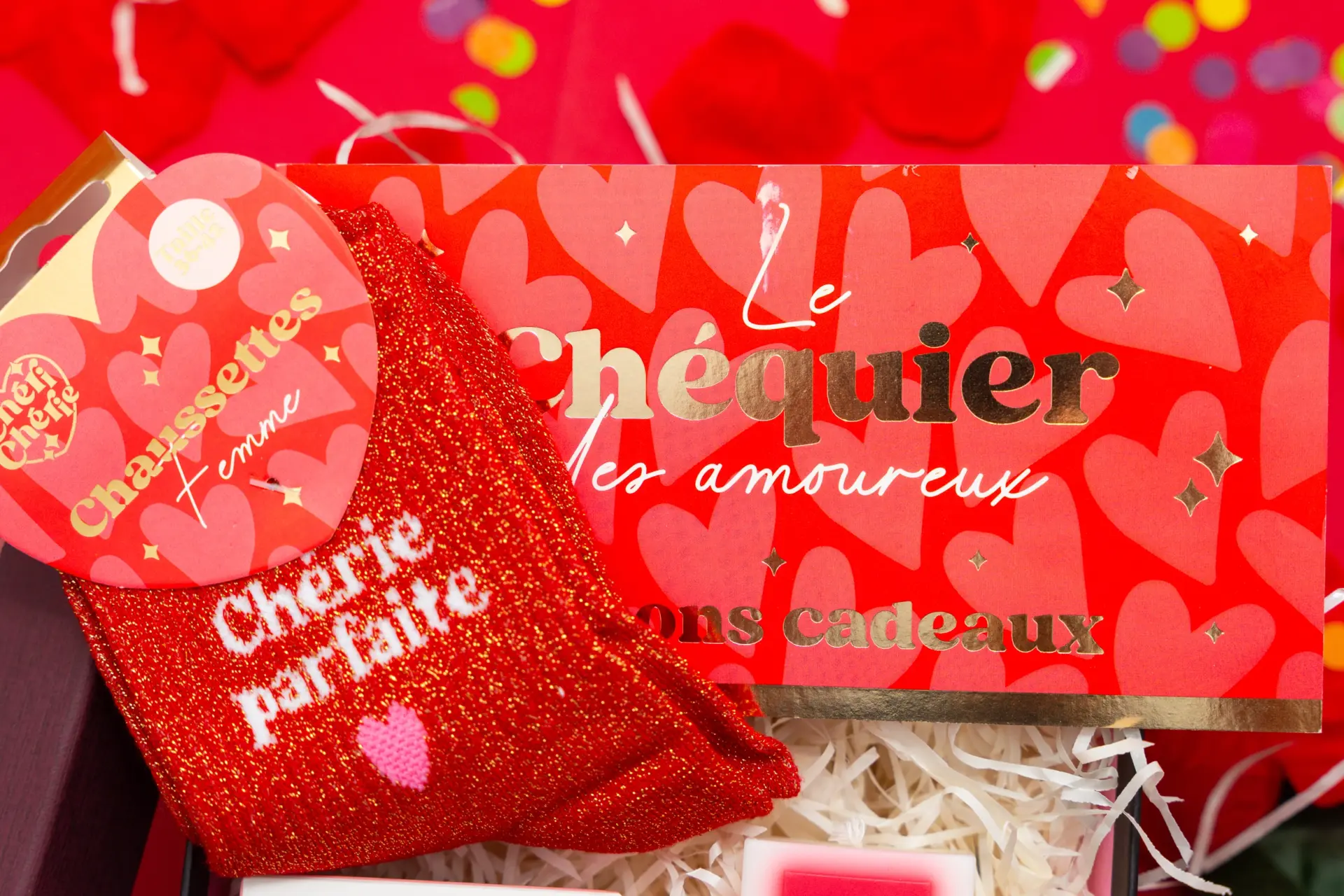 Love Box cadeau ouverte montrant des surprises romantiques : une paire de chaussettes rouges avec texte “Chérie parfaite”, un chéquier des amoureux avec motifs de cœurs à l’intérieur d’une boîte cadeau élégante.