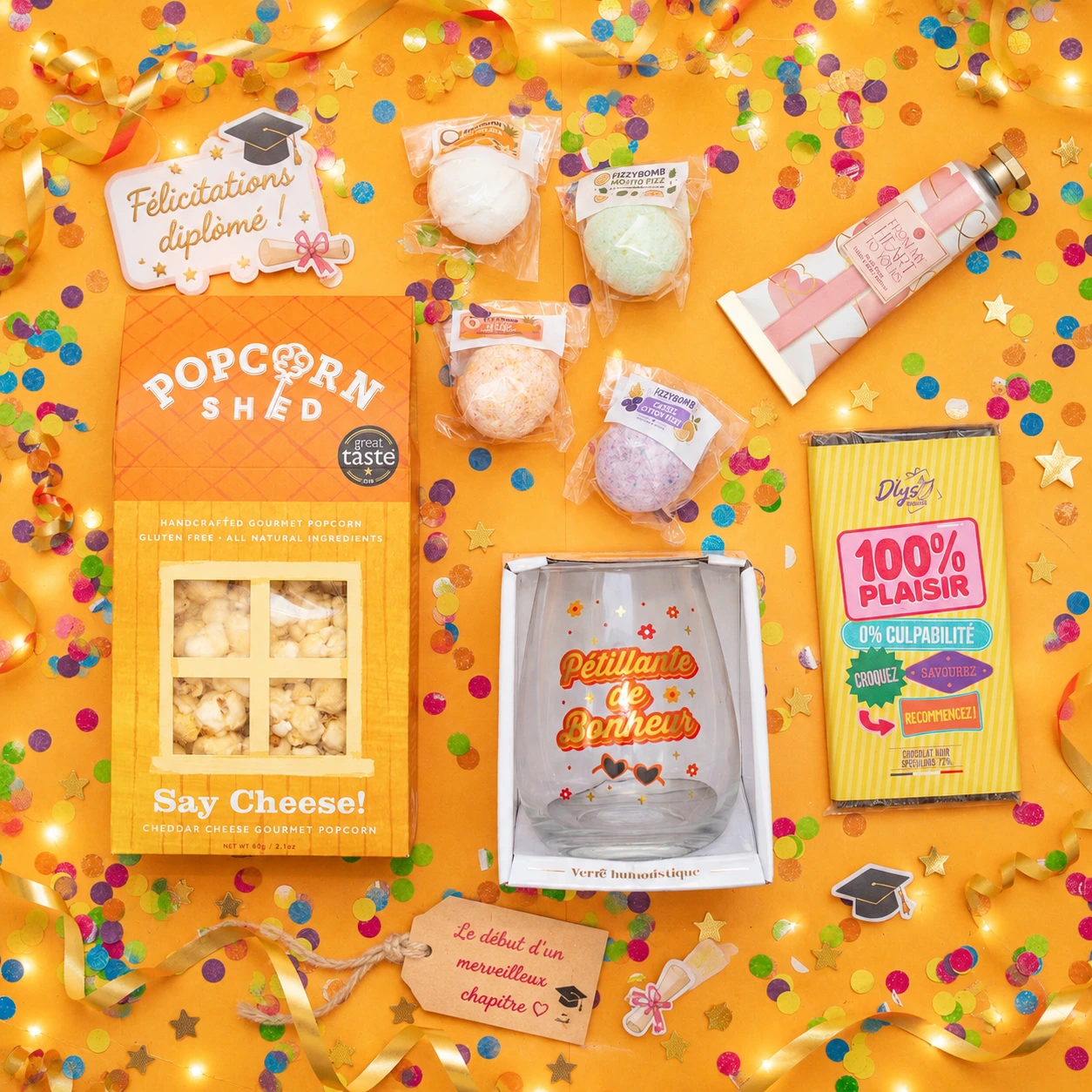 Vue de dessus du coffret diplôme personnalisable : popcorn cheddar, bombes cocktail sans alcool, verre humoristique, chocolat et crème mains sur fond jaune confettis