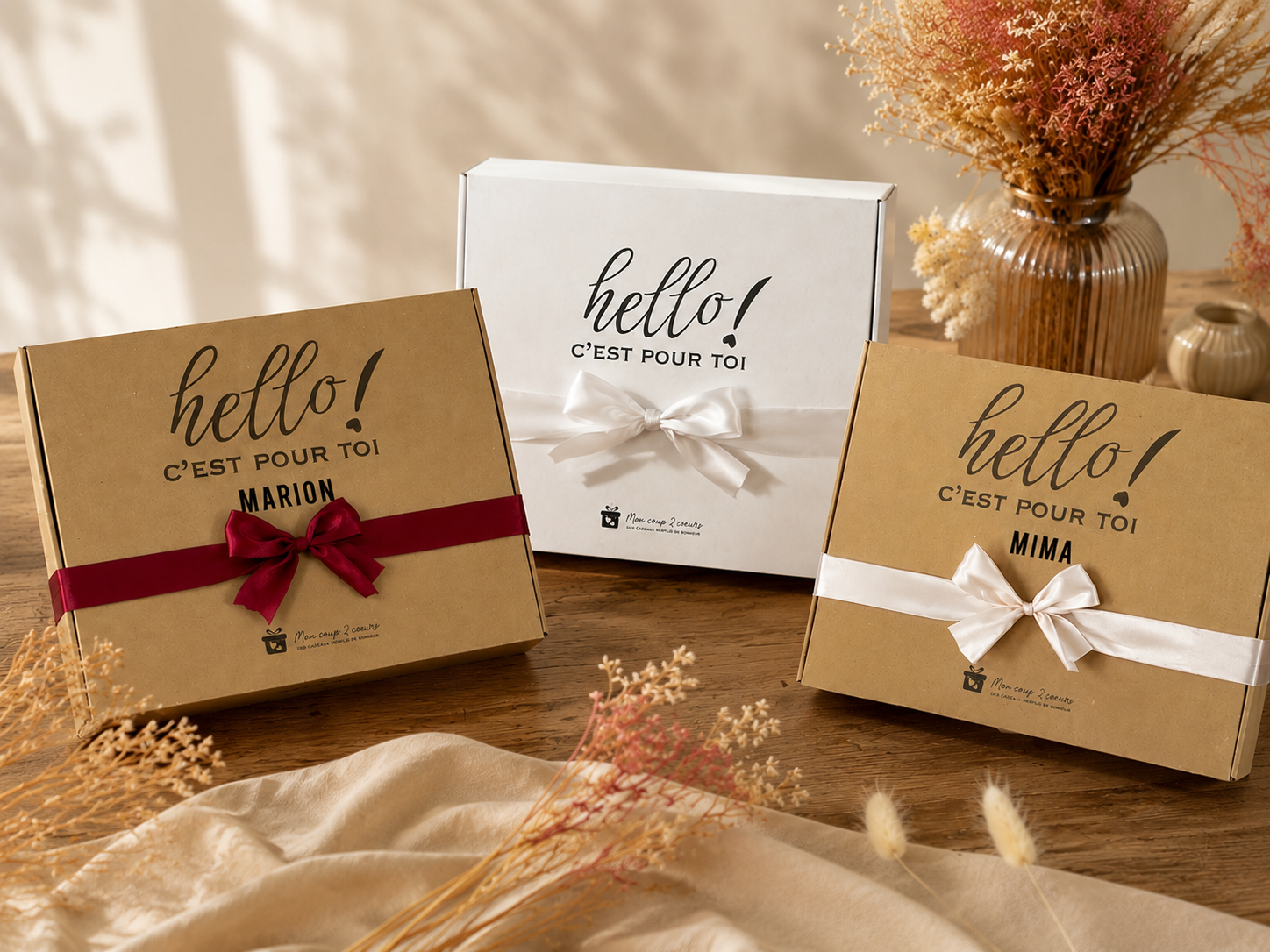 Trois coffrets cadeaux personnalisés avec nœuds en ruban et inscriptions « hello! C'est pour toi » de la marque Mon coup 2 cœurs sur une table en bois.